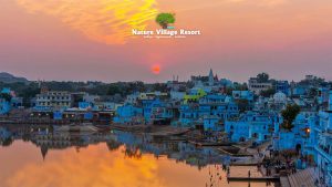A panoramic sunset view of the blue-painted houses and ghats of Pushkar, India, reflected in the still waters of Pushkar Lake. A red sun hangs low over the distant hills, under a warm orange and yellow gradient sky. The "Nature Village Resort" logo with a tree and sun graphic is centered in the upper sky, with text "Nature Village Resort relax . rejuvenate . restore".